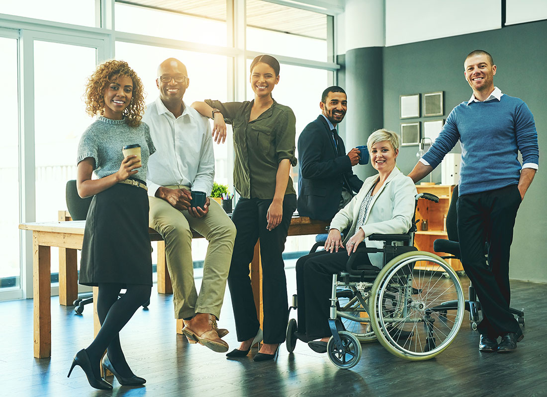 Employee Benefits - Group of Diverse Cheerful Employees Standing Next to and Sitting on a Desk in a Bright Modern Office Space with One Senior Employee in a Wheelchair
