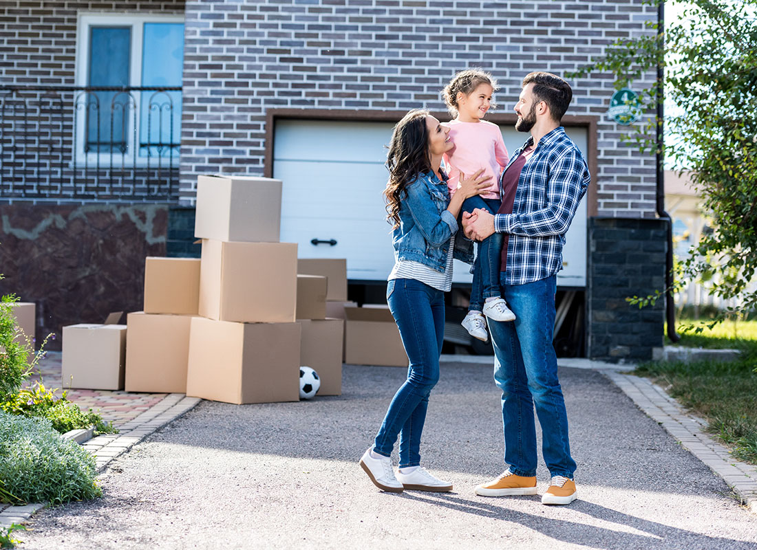 Personal Insurance - Portrait of Parents Holding Up Their Daughter While Standing Outside Their New Home in the Driveway with Stacks of Moving Boxes in Front of the Garage